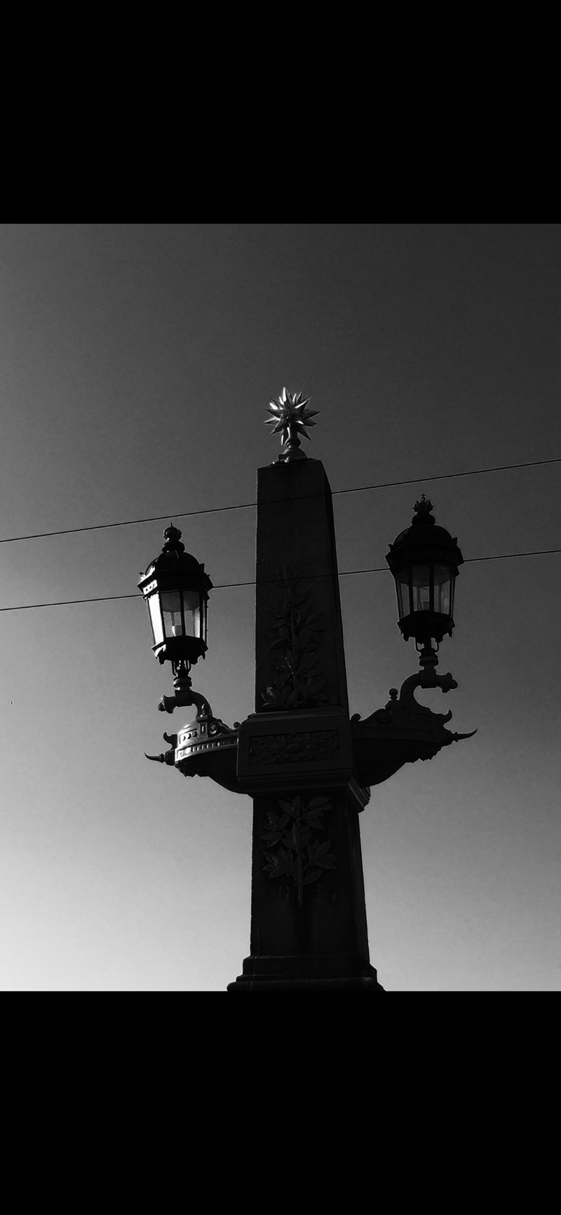 Black and white image of a tall, ornate streetlamp located on a bridge. The lamp post has two lanterns protruding from either side and a decorative star-shaped finial atop. Power lines run horizontally in the background against the sky, which transitions from light to dark.