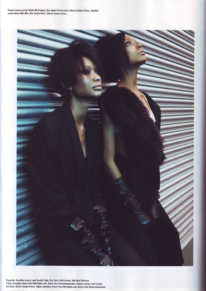 Two models pose in front of a horizontal corrugated metal backdrop. Both wear dark, sophisticated outfits with gloves. One model leans on the other with eyes closed, wearing a fur stole. The other looks off to the side with an intense gaze. The lighting casts soft shadows, enhancing the texture.