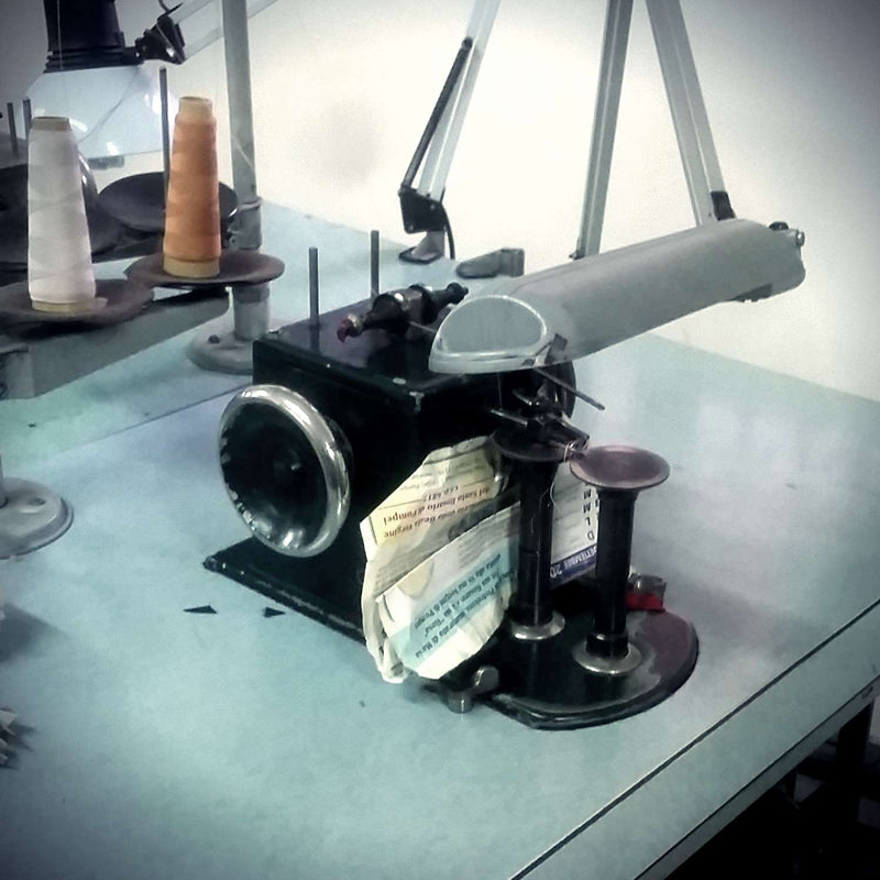 A vintage manual sewing machine is positioned on a gray table. The machine is black with a fabric guide and thread spool holders. It holds old, worn-out paper notes. Orange and beige thread spools are visible in the background, resting on a nearby shelf.