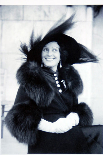 Black and white photo of a smiling woman dressed in an elegant outfit. She is wearing a large, wide-brimmed hat with feathers, a fur-trimmed coat, and white gloves. Her earrings are long and decorative. The background is neutral, drawing focus to her sophisticated attire and cheerful expression.