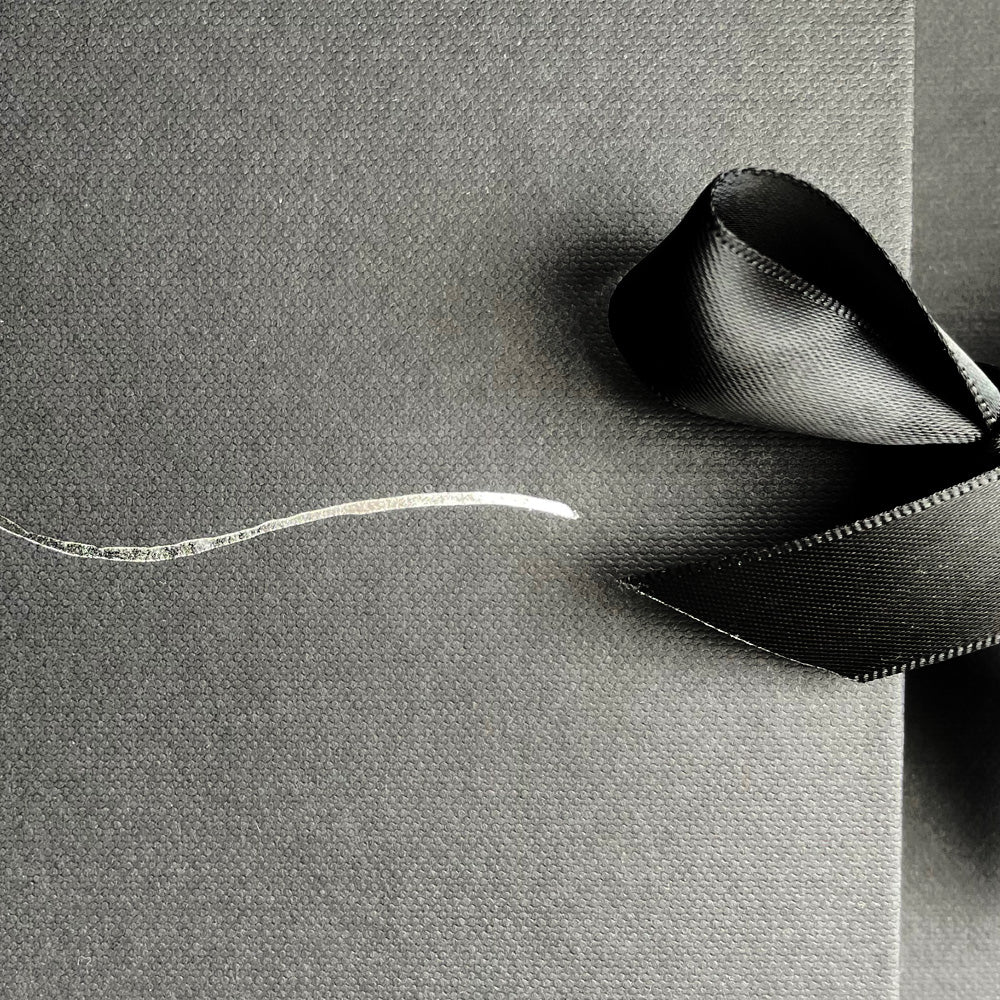 A close-up image of black textured paper, partially covered by a smooth, glossy black ribbon tied into a simple loop. A thin, slightly curved, silver metallic line decorates the left side of the paper, adding a subtle contrast to the dark, elegant background—a perfect setting for an Ines Gloves Gift Card from Ines Gloves.