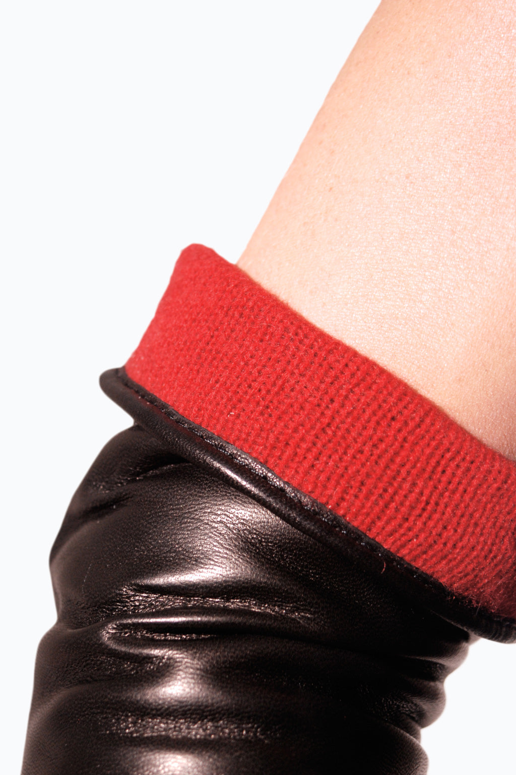Close-up of a person's wrist wearing Ines Gloves' Long cashmere lined gloves with a red knit cuff. The image focuses on the contrast between the shiny, smooth texture of the black leather and the soft, textured red knit. The long gloves feature an elegant opera length design. A plain white background makes them stand out.
