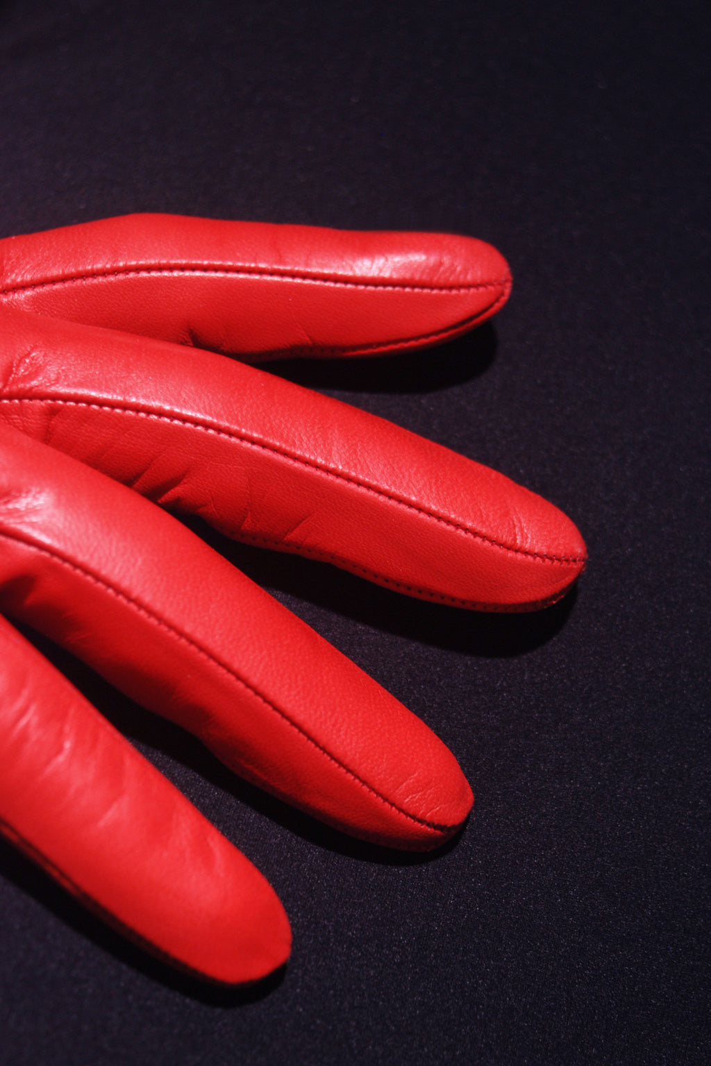 A close-up image of **Ines Gloves Long cashmere lined gloves** placed on a dark, possibly black surface. The lighting accentuates the texture and seam details of the gloves, revealing their luxurious cashmere lining. Only the fingers of the glove are visible, fanned out slightly. The contrast between the vibrant red and the dark background is striking.